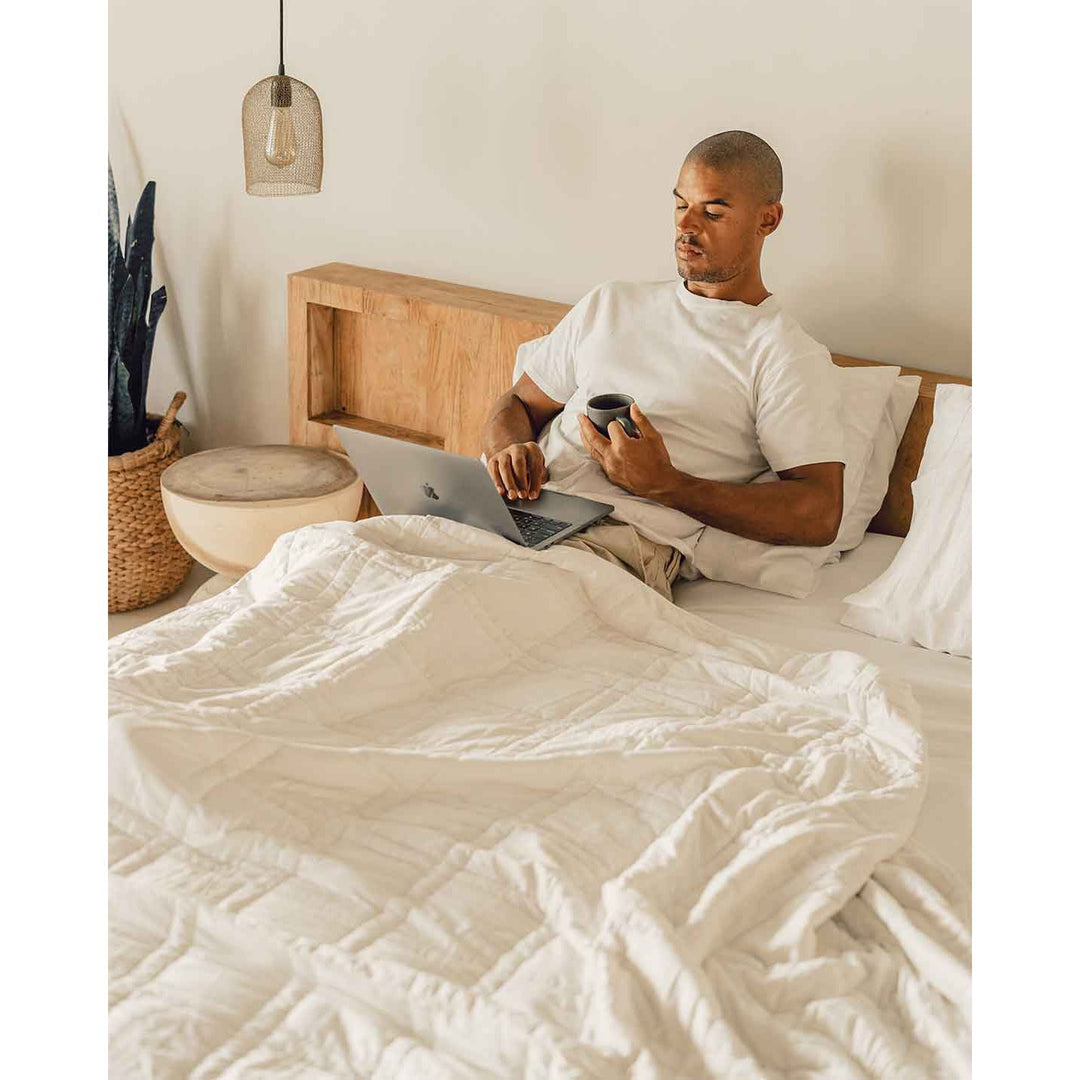 A man with a shaved head holding a cup of coffee sits up in bed with a laptop, under a weighted comforter.  [32783560769616, 32783560802384, 32783560933456]