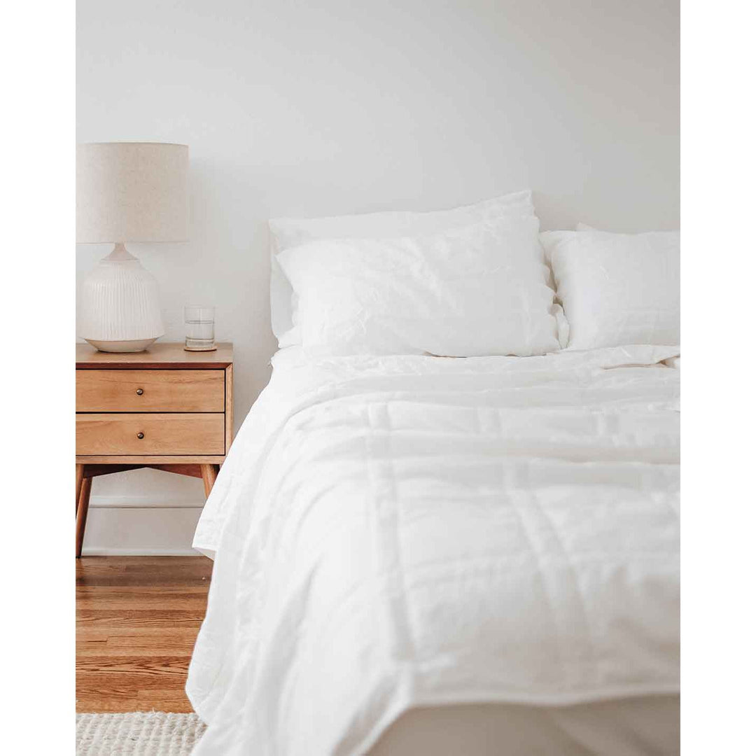 The side of a bed with a white comforter rumpled, white pillows and bedside table with a glass of water . [32783560769616, 32783560802384, 32783560933456]