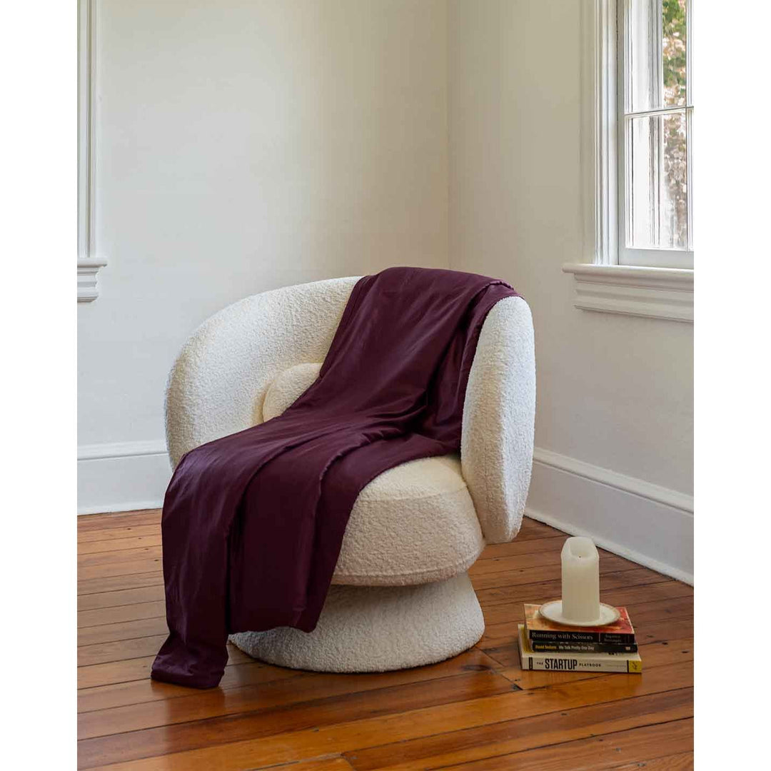 A mulberry cotton duvet cover is tossed over a chair next to a candle and a stack of books [44422682149083]