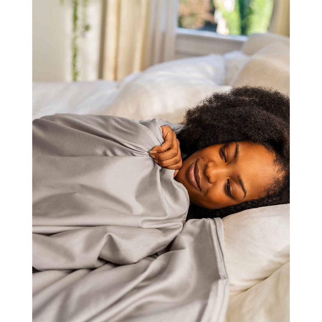 A woman looks down while relaxing in bed holding a cotton duvet cover up to her chin [44422682050779, 45027099771099]