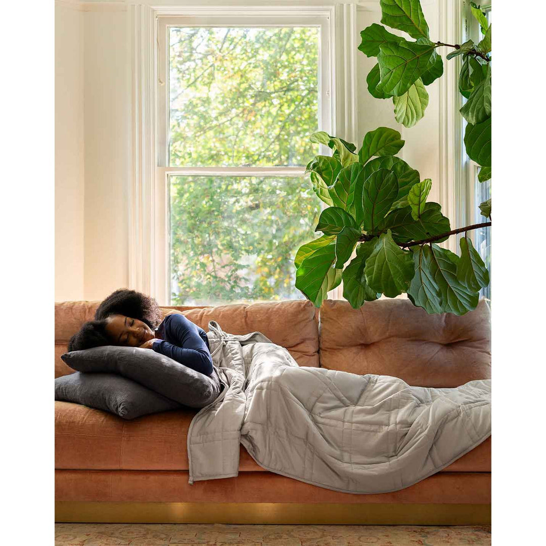 A woman in blue shirt naps under a silver sage weighted blanket on a pink sofa with a fig tree [42952280801499, 43631556591835, 43631556952283, 43631557968091]