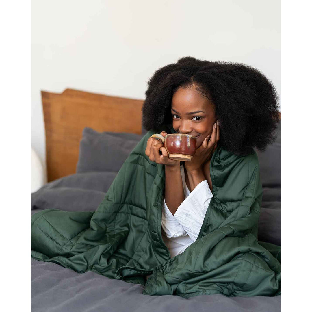 A woman smiles and takes a sip of coffee with a spruce green weighted blanket wrapped around her shoulders [44422820561115, 44422814826715, 44422813090011, 44422818234587, 45574418333915, 45574418235611, 45574418202843, 45574418235611, 45574418202843, 45574418301147]