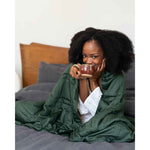 Load image into Gallery viewer, A woman smiles and takes a sip of coffee with a spruce green weighted blanket wrapped around her shoulders [46061921730779, 46061921763547, 46061921796315, 46063267709147, 46063269118171]
