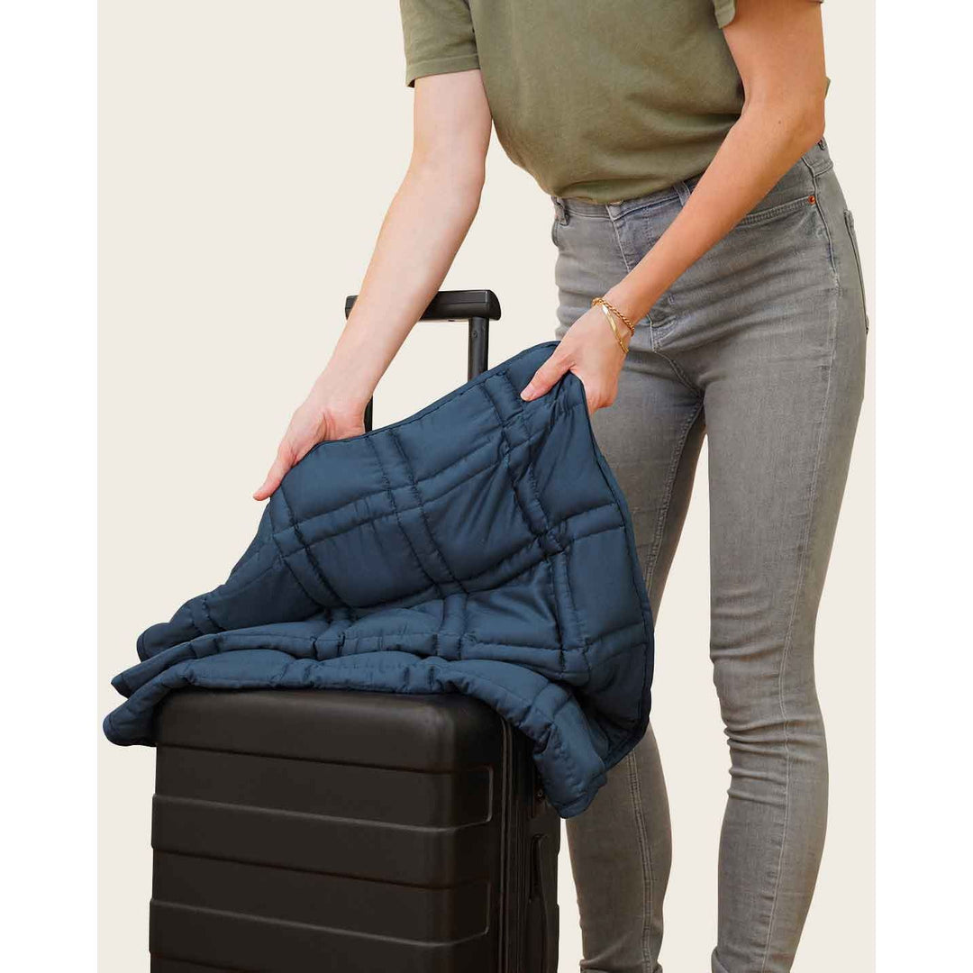 A woman holds a luna blue lap blanket with her suitcase, showing that she is taking it to travel. [4358065671394-7, 4628803738031-5]