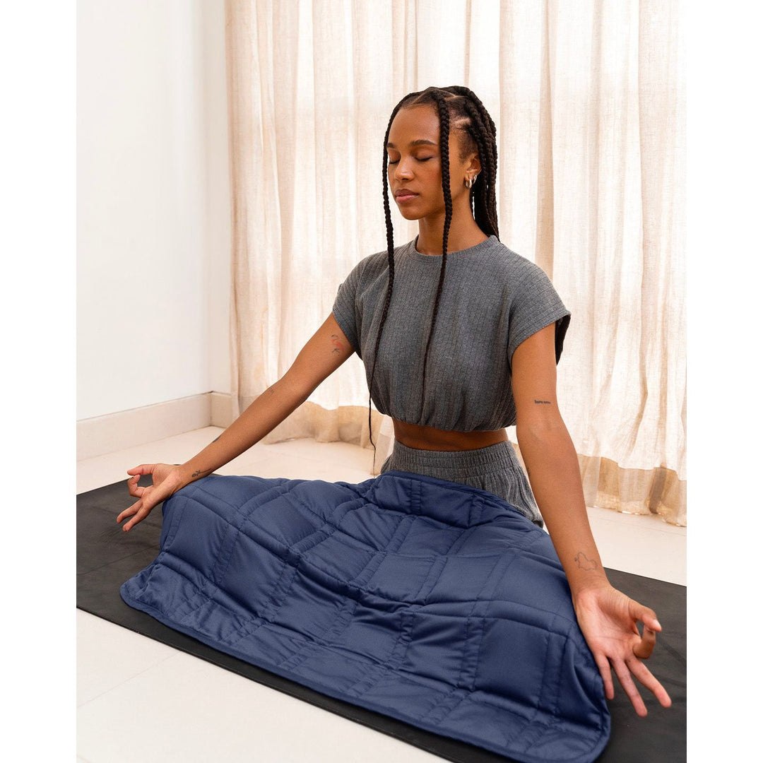 A young woman with braided hair sits cross legged on a yoga mat with her eyes closed. She has a luna blue lap blanket across her lap while she meditates. [43580656713947,46288037380315]