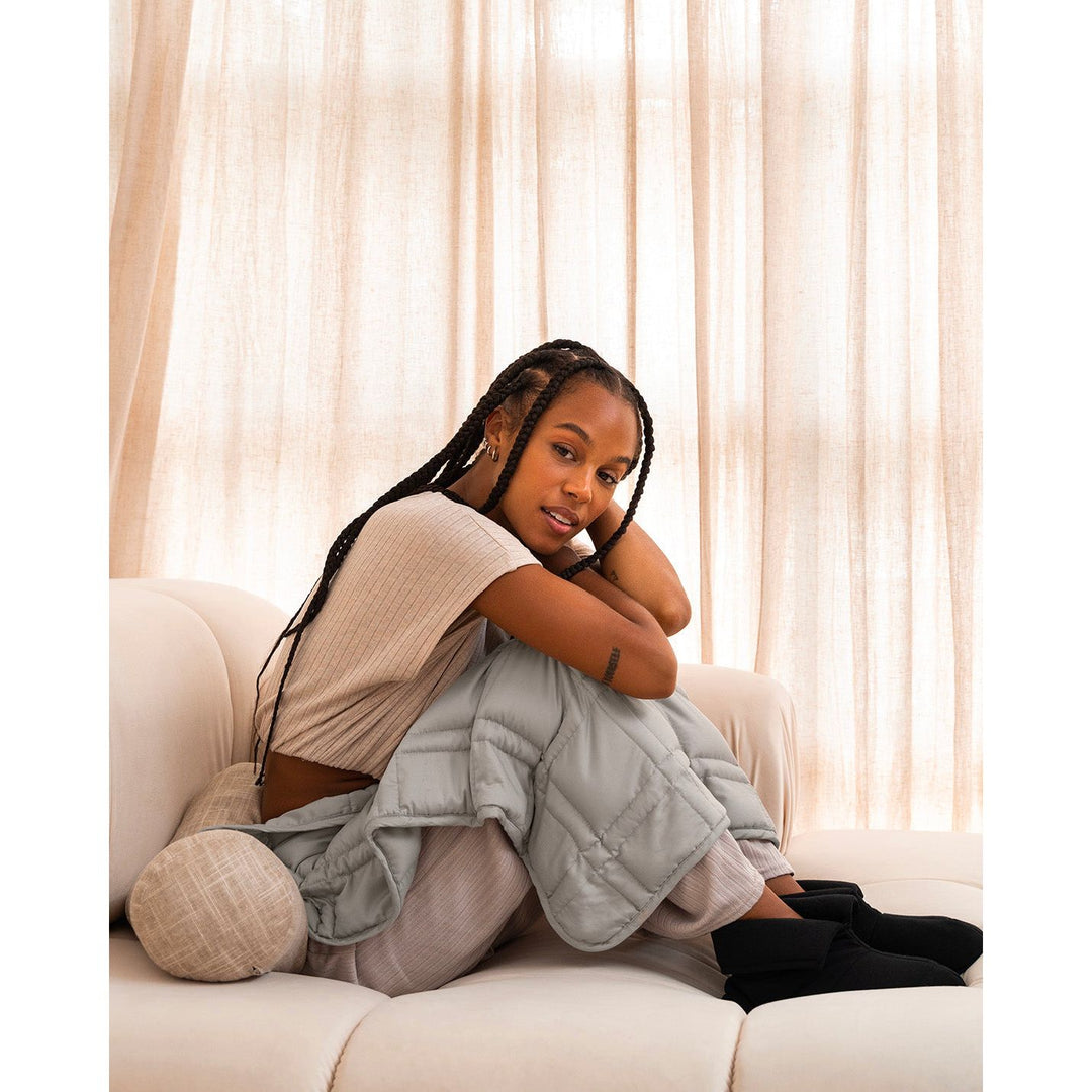 A young woman with braids sits on a beige sofa in loungwear, with a silver sage lap blanket on her lap. [43580656746715, 46288037413083]