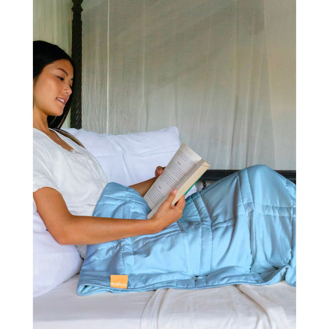 A young woman reads in bed with the indigo weighted blanket on her lap [44385363722459]