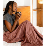 Load image into Gallery viewer, [44676999741659] woman holding a cup and wearing a weighted blanket in bed. 
