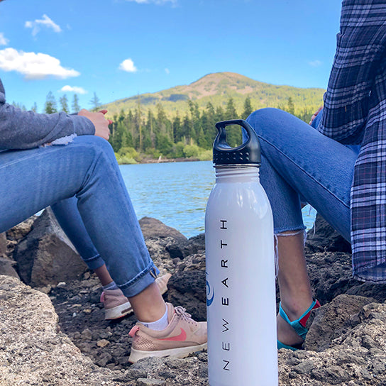 A white New Earth water bottle sitting on a rock next to two woman who are enjoying the lake view. 