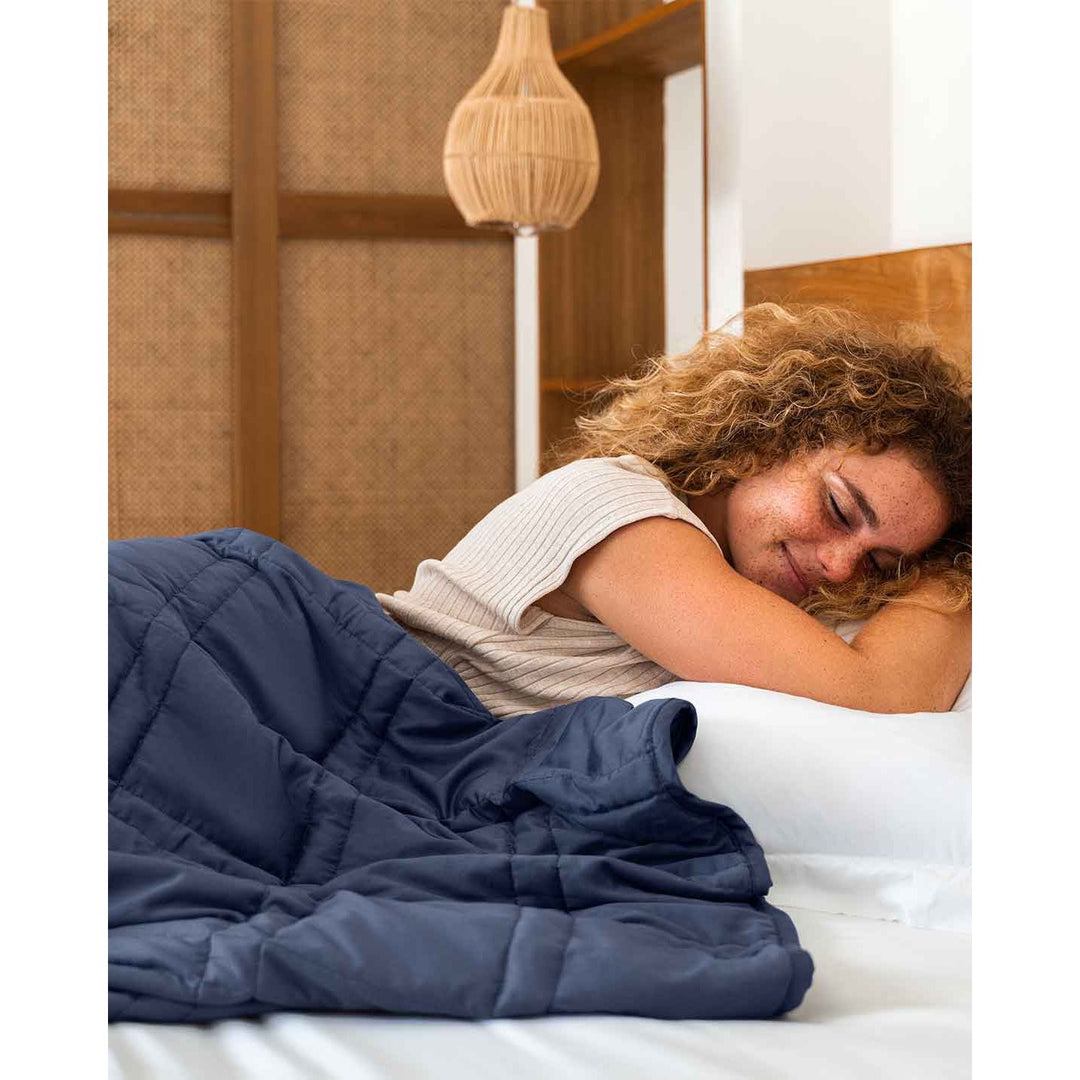 A young woman with red curly hair lies in a bed with a blue weighted blanket across her body. [43649942782171, 44422804340955, 44422811287771, 44422816432347]