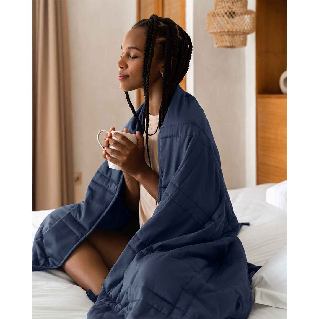 A young woman sits cross legged in a bed with a blue weighted blanket wrapped around her shoulders, and holds a warm cup of coffee. [44385233109211,46089486074075]