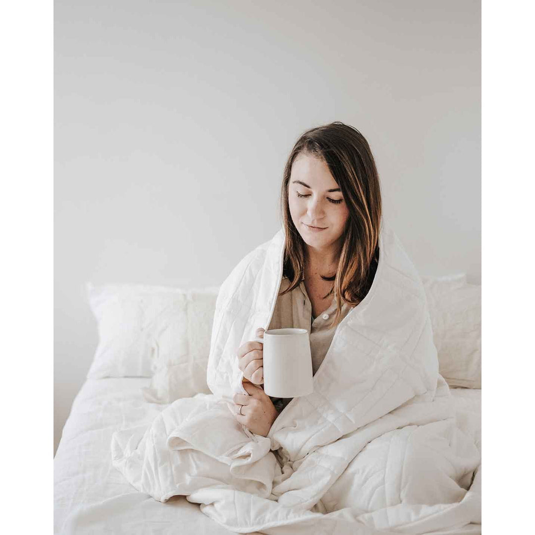 A young woman with brown hair sits cross legged on a bed with a white weighted blanket wrapped around her shoulders and holding a mug, softly smiling. [44385233043675]