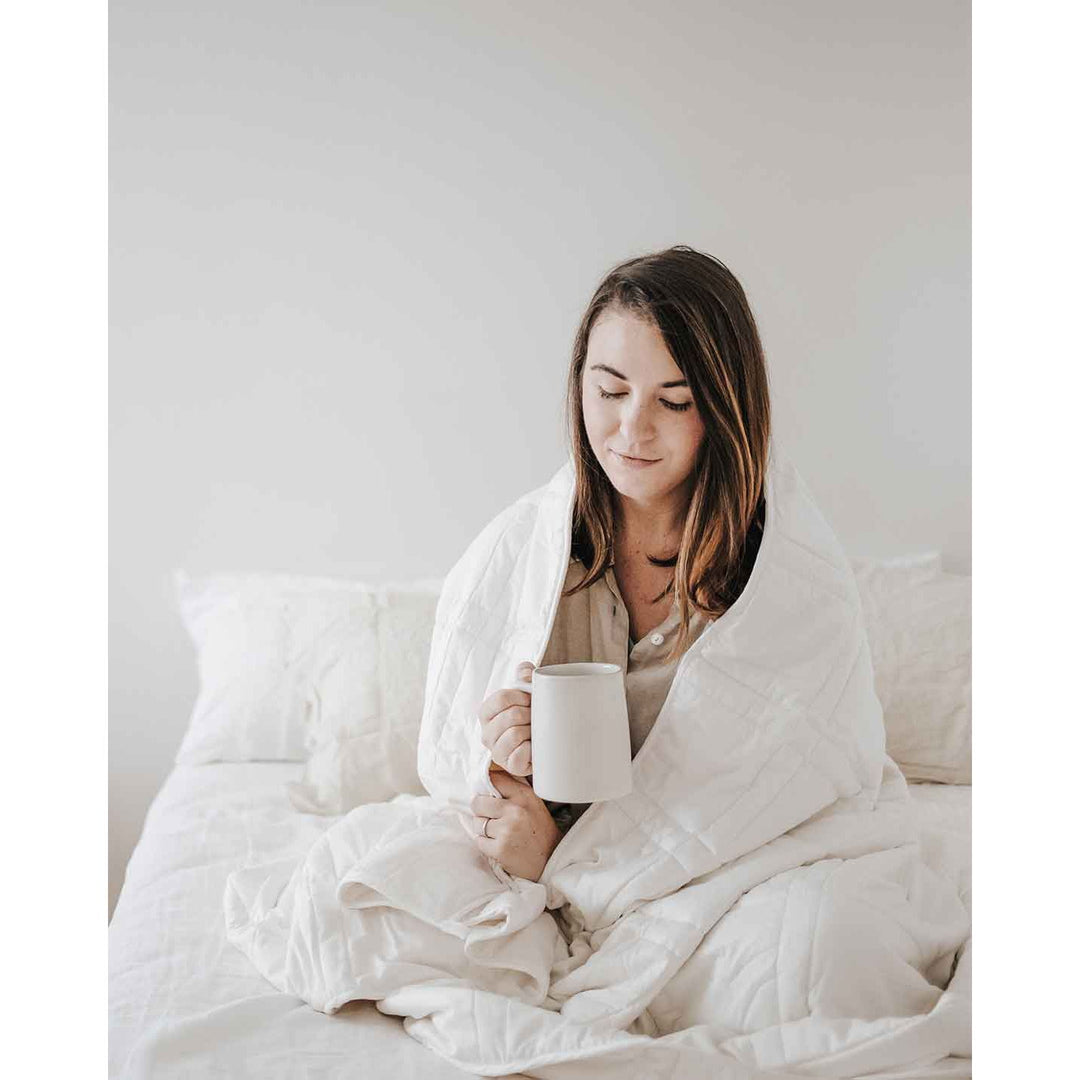 A young woman with brown hair sits cross legged on a bed with a white weighted blanket wrapped around her shoulders and holding a mug, softly smiling. [42934587621595, 45574417809627]