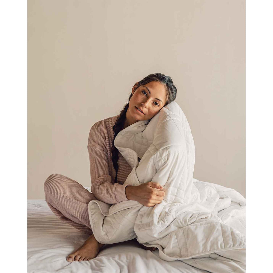 A young woman sits cross legged on a bed wearing soft pink pajamas and holding a white weighted blanket to her face. [44385233141979, 44385233207515, 44385233273051]