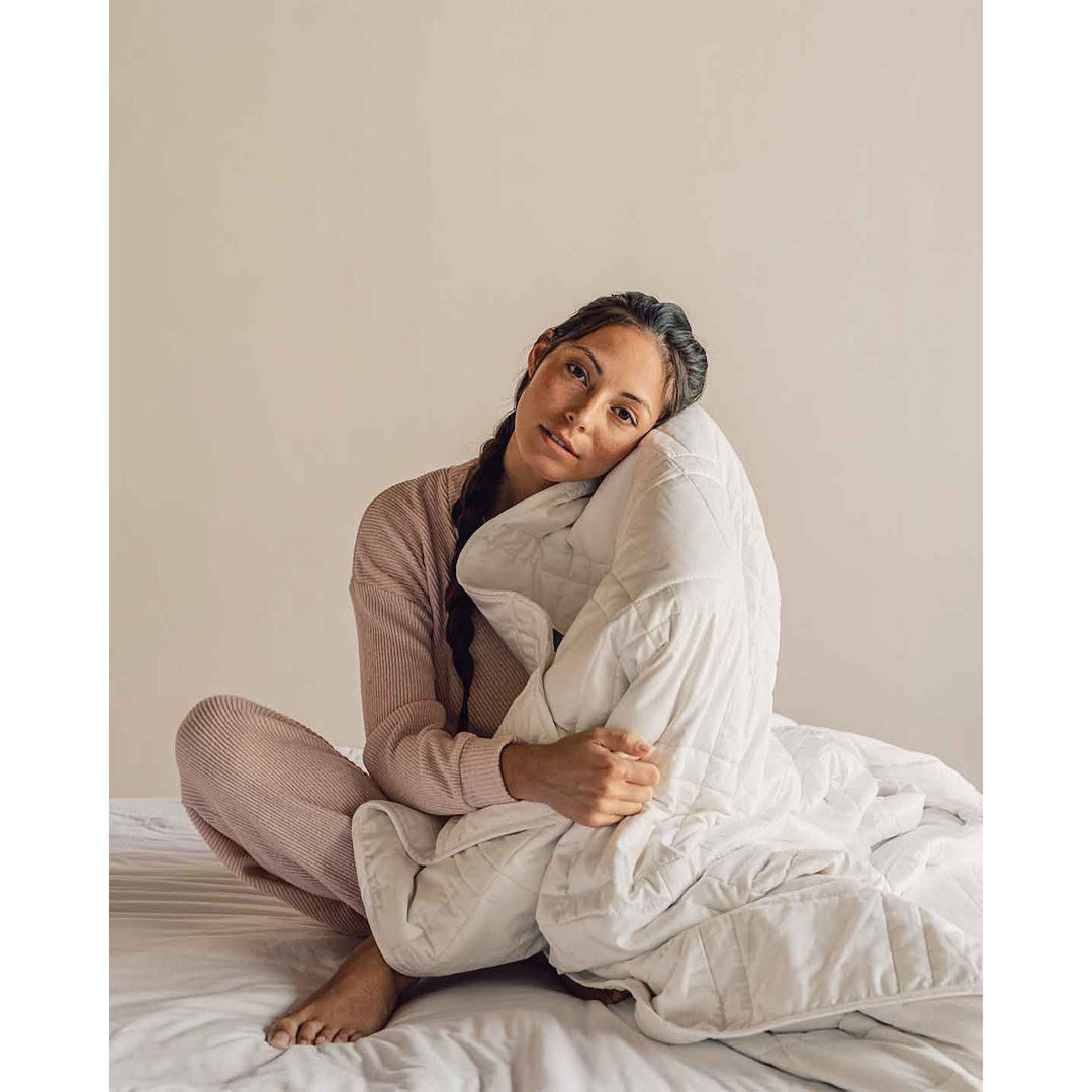 A young woman sits cross legged on a bed wearing soft pink pajamas and holding a white weighted blanket to her face. [42934587621595, 45574417809627,42934587883739, 46452819001563]