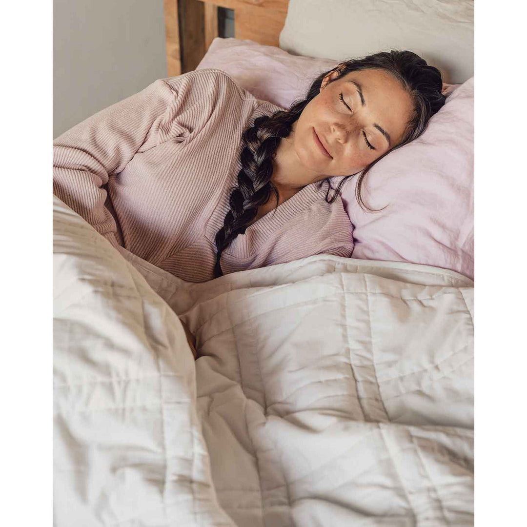 A young woman with a braid sleeps peacefully in a bed with a pink pillowcase and a white weighted blanket across her body. [44385233043675]