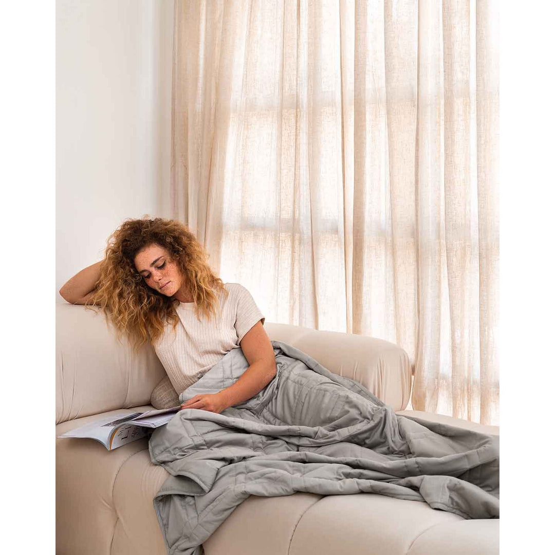 A young woman with curly red hair and freckles rests on a cream sofa reading a magazine, with a silver sage weighted blanket across her body. [44385233076443, 44385233174747, 44385233240283, 44385233305819]