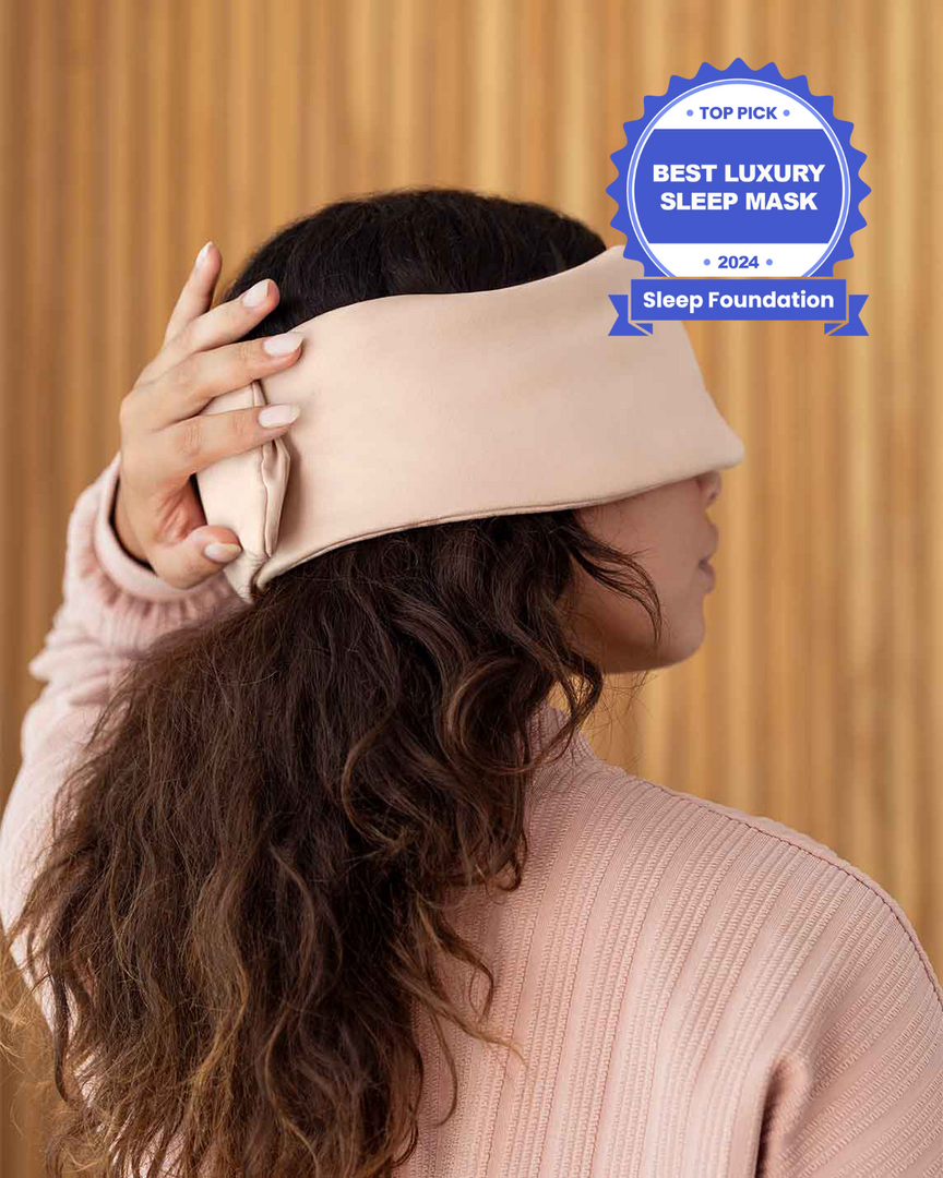 A photo from the side of a woman with long brown hair holding her hand over the back of a silk sleep mask. [44377020825819, 46730820944091, 46730821009627, 46730821075163, 46730821140699]