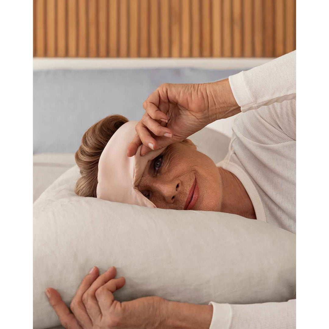 A senior woman with blonde hair lays down on a pillow wearing a pink silk sleep mask, holding one side up to view the camera [44377020825819]