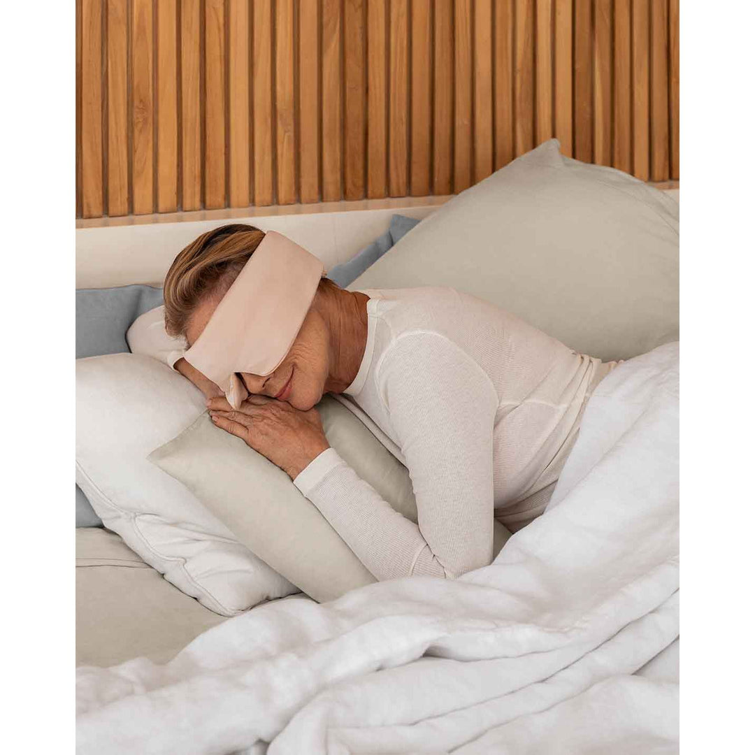 A senior woman with blonde hair rests her head on a pillow wearing a silk sleep mask [44377020825819]