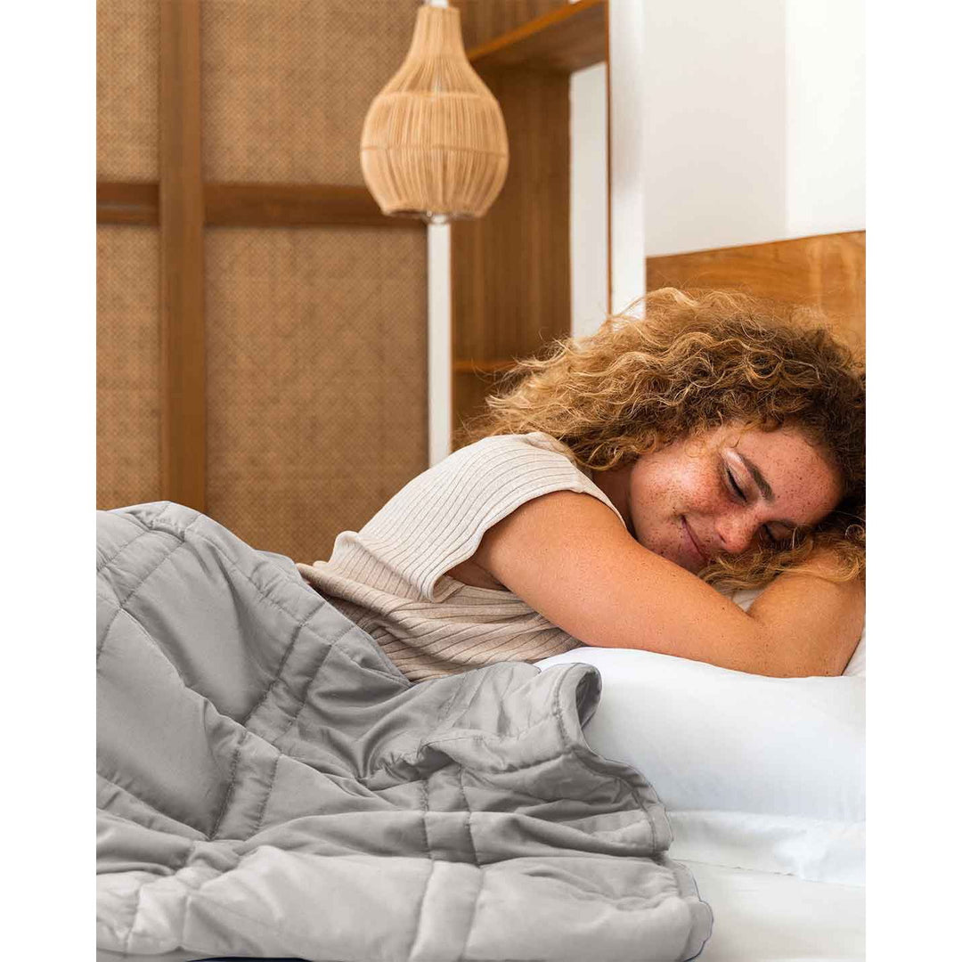 A young woman with curly red hair and freckles lies down in a bed with a silver sage weighted blanket. [42952280801499, 43631556591835, 43631556952283, 43631557968091]